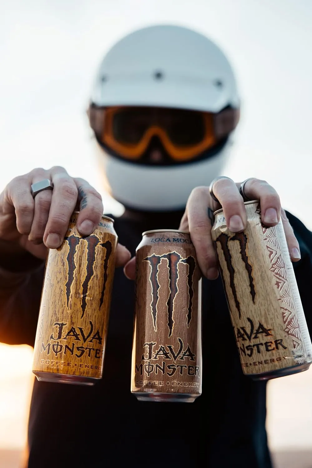 A person wearing a white helmet and orange-tinted goggles holds three tall Java Monster cans toward the camera