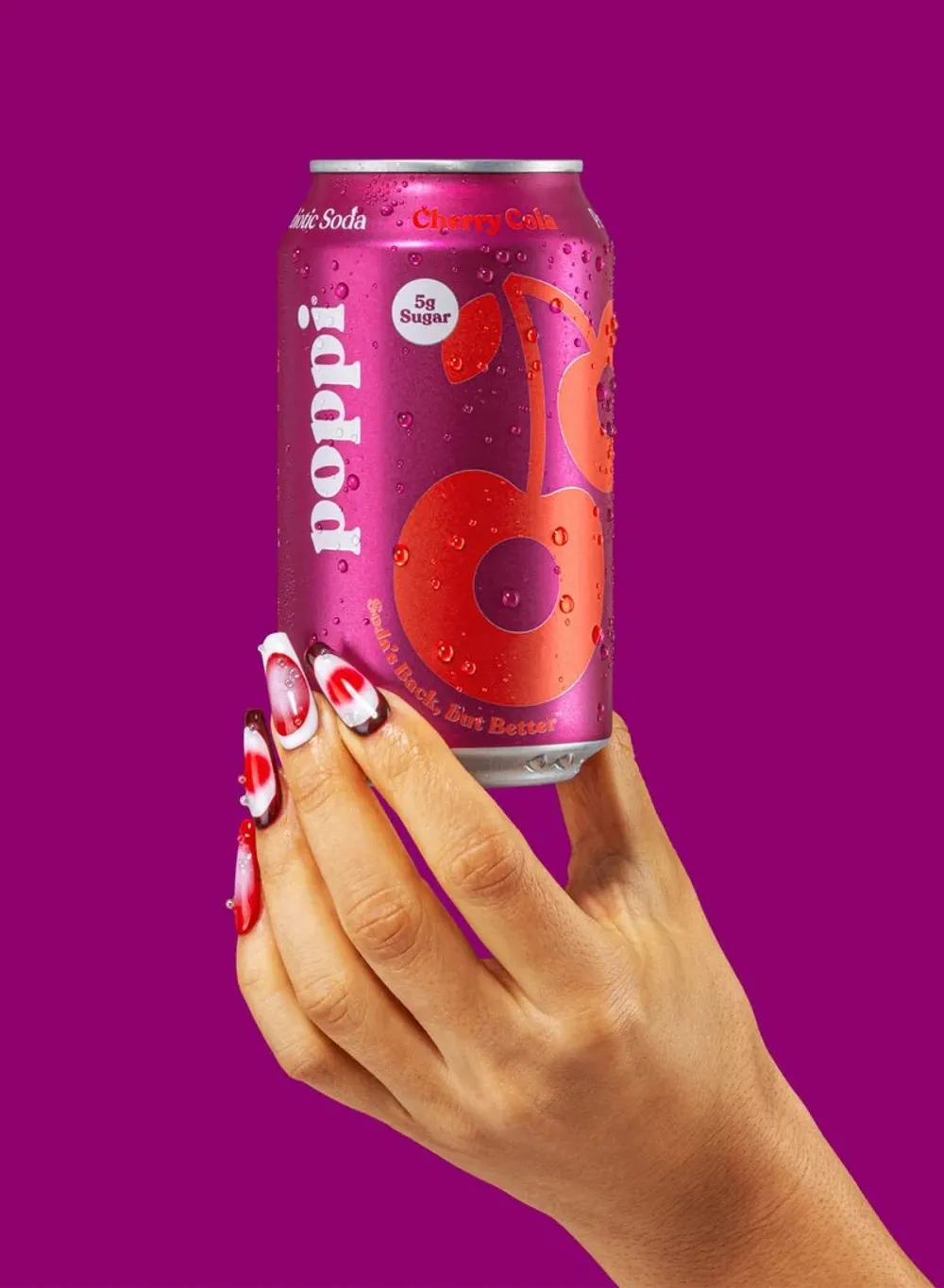 A hand with red-and-white decorated nails holds a pink Poppi can against a solid magenta background