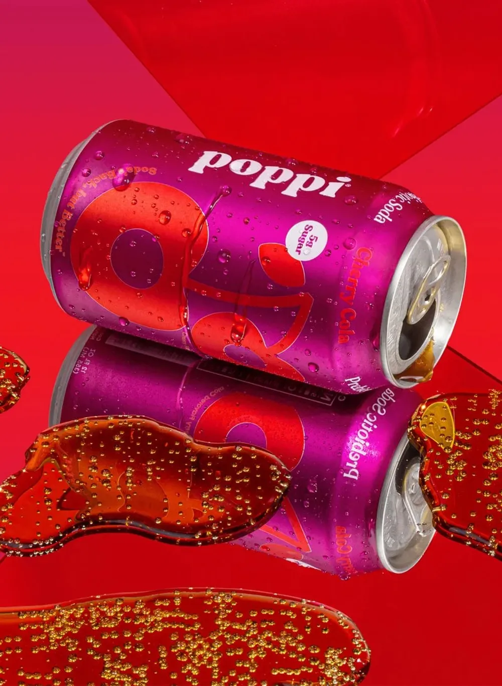 Two chilled Poppi soda cans lie horizontally against a bright red background, with condensation and splashes of dark red liquid around them
