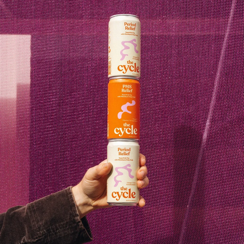 A hand holds three stacked cylindrical The Cycle drink containers against a textured magenta backdrop